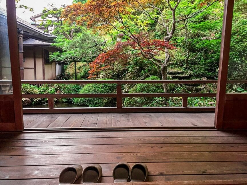 Three pairs of sandals placed on a wooden deck overlook a serene Japanese garden in Koyasan, with lush greenery and colorful foliage in the background, evoking a sense of ancient Japanese culture.