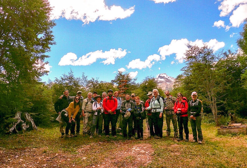 A group of hikers in outdoor gear stands together in a clearing surrounded by trees, with a mountain visible in the background under a partly cloudy sky, ready to embark on one of Patagonia's epic hikes.