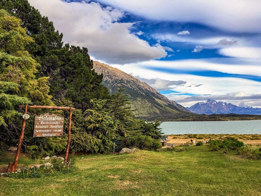 Wooden sign on a grassy area near a lake with mountains in the background. Pine trees and bushes surround the area under a partly cloudy sky, reminiscent of Patagonia's epic hikes or vistas found in National Parks.