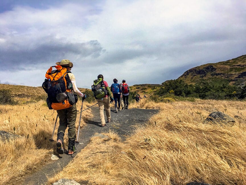 A group of hikers with backpacks walks along a dirt trail through a grassy landscape under a cloudy sky, showcasing one of Patagonia's epic hikes.
