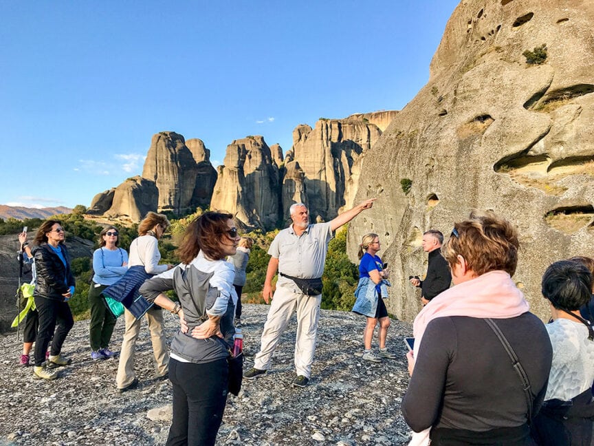 A tour guide gestures towards rock formations while a group of tourists listens and observes the natural landscape, reminiscent of the rugged beauty found on the nearby isles of Greece.