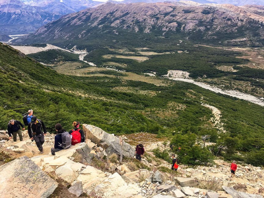 Hikers tackle a rocky trail in a mountainous landscape, with green valleys and a distant river below under a clear sky, reminiscent of the epic hikes found in Patagonia's famed National Parks.