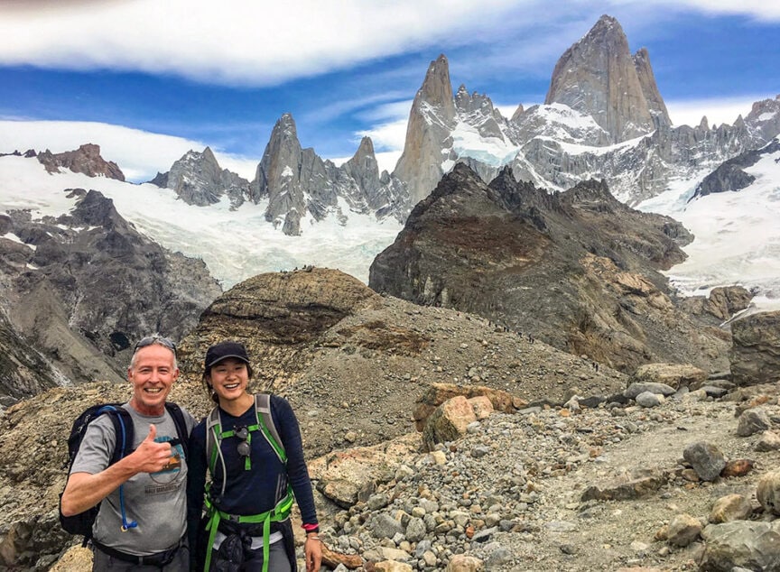 Two hikers smile and pose on a rocky trail in Patagonia, with towering, jagged snow-capped mountains in the background under a partly cloudy sky, capturing the essence of epic hikes found in national parks.
