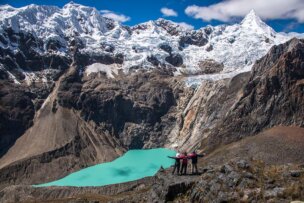 Two people stand on a rocky ledge with arms outstretched, celebrating a 60th birthday while overlooking a turquoise glacial lake surrounded by rugged mountains covered in snow under a blue sky.