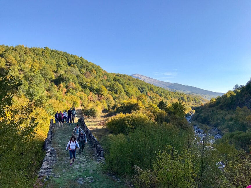 A group of people hikes on a trail through a lush, green hillside under a clear blue sky, exploring the scenic beauty beyond Athens.