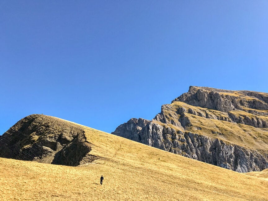 A lone person walks on grassy terrain with rocky hills and a clear blue sky in the background, evoking the spirit of exploring Greece.