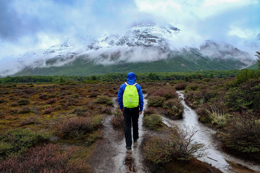 A person wearing a blue jacket and green backpack is hiking on a muddy trail toward a snow-capped mountain surrounded by mist and low clouds, embodying the spirit of adventure in Patagonia.