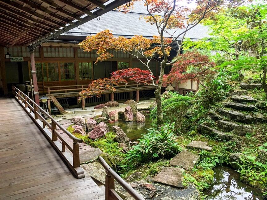 Traditional Japanese courtyard with a wooden walkway, a small pond, stone steps, and various plants and trees, including a Japanese maple with red leaves. Reflecting the essence of ancient Japanese culture, a wooden building is visible in the background.