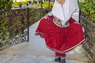 A young girl exudes beauty in traditional red clothing and a headscarf, posing outdoors near ornate iron gates in Iran, holding out her skirt with an air of warmth.