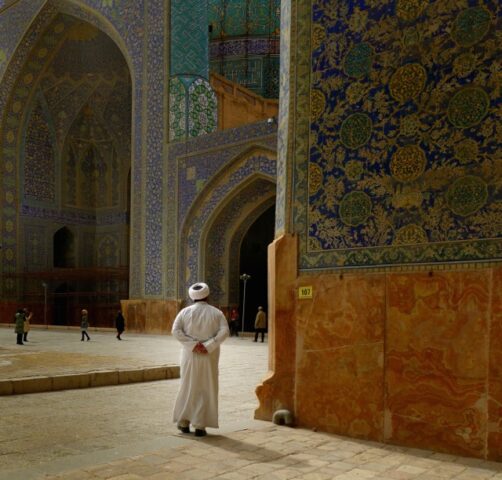 A person in traditional attire stands in front of ornate, tiled walls at a historical or religious site in Iran, radiating beauty and warmth while others walk in the background.