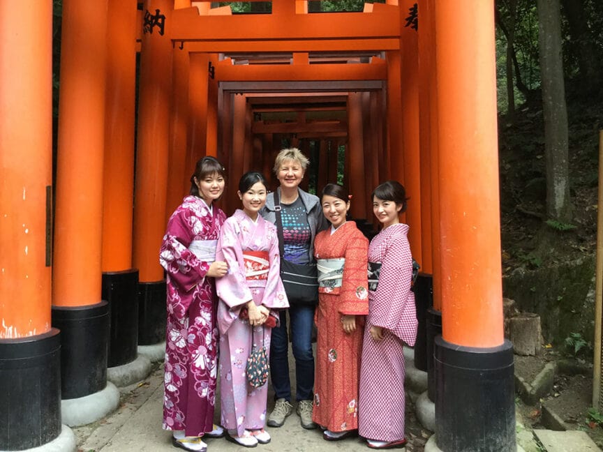 A group of five people, including four women in traditional Japanese kimonos and one person in casual attire, standing in front of red torii gates on a historic hiking trail in Shikoku.