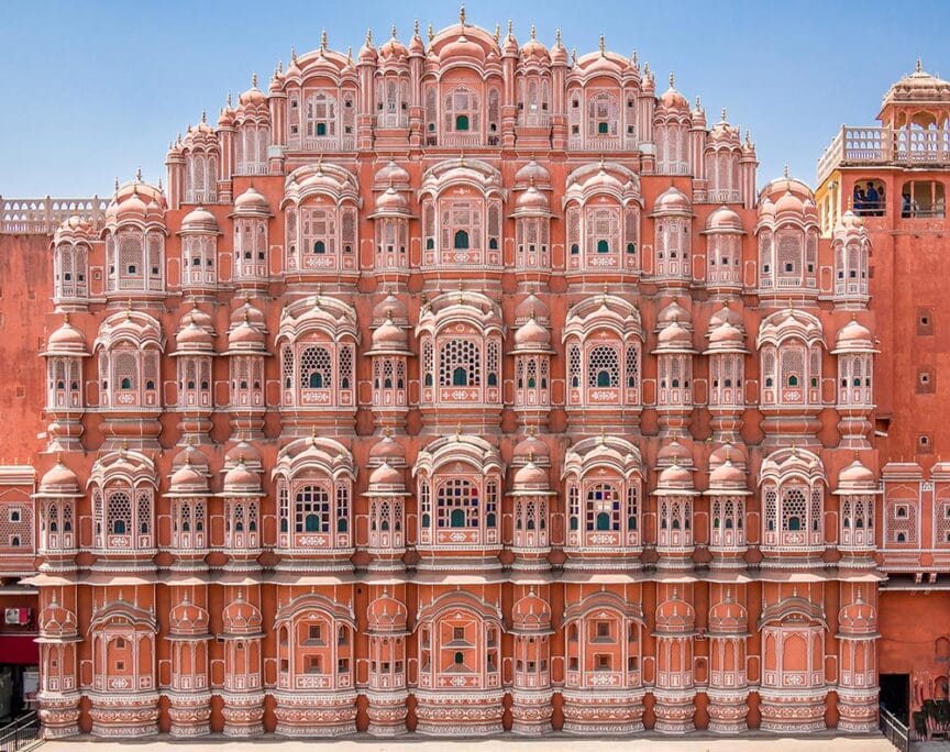 A grand, symmetrical pink sandstone building with numerous small windows and intricate latticework, the Hawa Mahal stands proudly in the vibrant destination of Jaipur, India, under a clear blue sky.