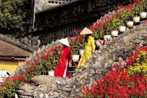 Two individuals wearing traditional conical hats and colorful attire ascend a dragon-shaped stone staircase adorned with vibrant red flowers and white potted plants, capturing the essence of Adventures in Indochine.
