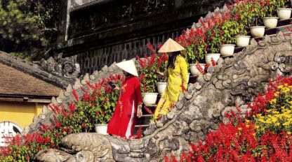Two individuals wearing traditional conical hats and colorful attire ascend a dragon-shaped stone staircase adorned with vibrant red flowers and white potted plants, capturing the essence of Adventures in Indochine.