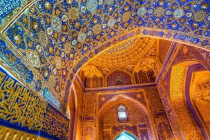 A detailed view of a mosque's ornate interior showcases intricate blue and gold geometric patterns and Arabic calligraphy on its arched ceiling and walls, illuminated by soft, warm light, capturing the essence of a Silk Road cultural journey.