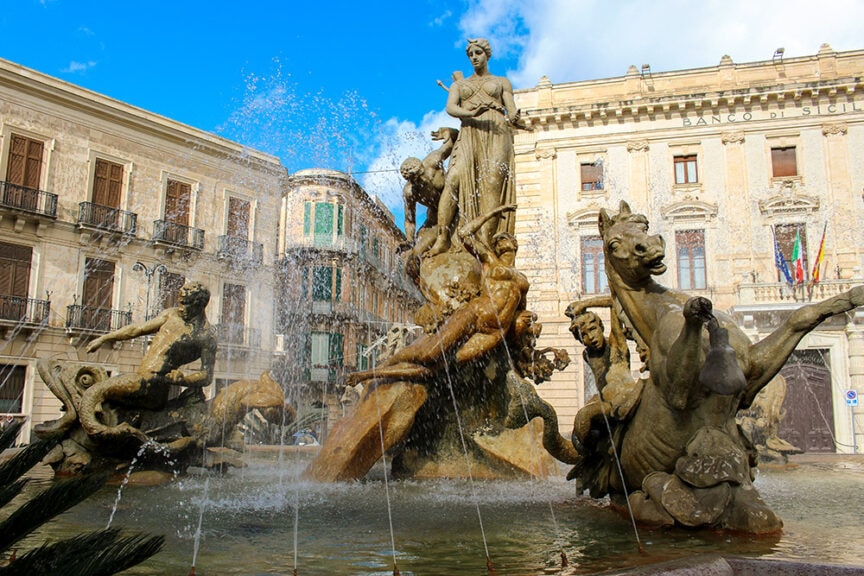 Fountain with multiple statues depicting mythological figures and creatures, set in a historic city square surrounded by classical buildings, embodying an essence of urban antiquity akin to the ancient allure of Petra or historic charm found in Peru.