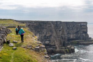 Three hikers with backpacks walk along a grassy cliff edge with steep rocky faces overlooking the ocean on a cloudy day, exploring Ireland's West Coast.