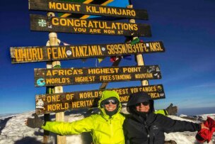 A grandmother and mother in winter gear stand in front of a sign at the summit of Mount Kilimanjaro, which reads: "Congratulations. You are now at Uhuru Peak, Tanzania, 5895m/19341ft ASL, Africa's Highest Point." Their Climbing Kili adventure has brought three generations to this breathtaking peak.