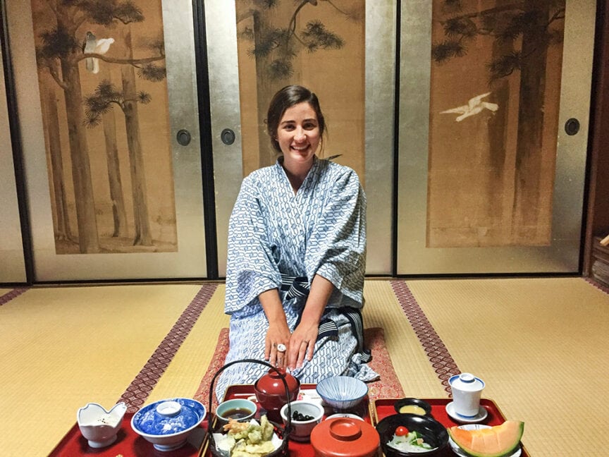 A woman in a traditional robe sits on a tatami mat with a Japanese meal arranged in front of her, embodying ancient Japanese culture. Behind her, sliding doors adorned with painted trees evoke the serene ambiance of Koyasan.