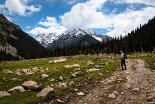 A hiker walks on a rocky path through a grassy field with scattered rocks, surrounded by tall trees, with a view of snow-capped mountains under a partly cloudy sky in the background. The scene offers peaceful reflections reminiscent of a family trip in Kyrgyzstan.