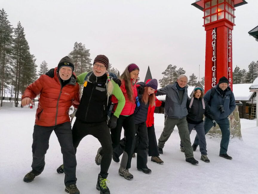 A group of eight people dressed in winter clothing pose together outdoors near a red sign reading "Arctic Circle" in a snowy, wooded area, capturing the essence of Northern Finland adventures.