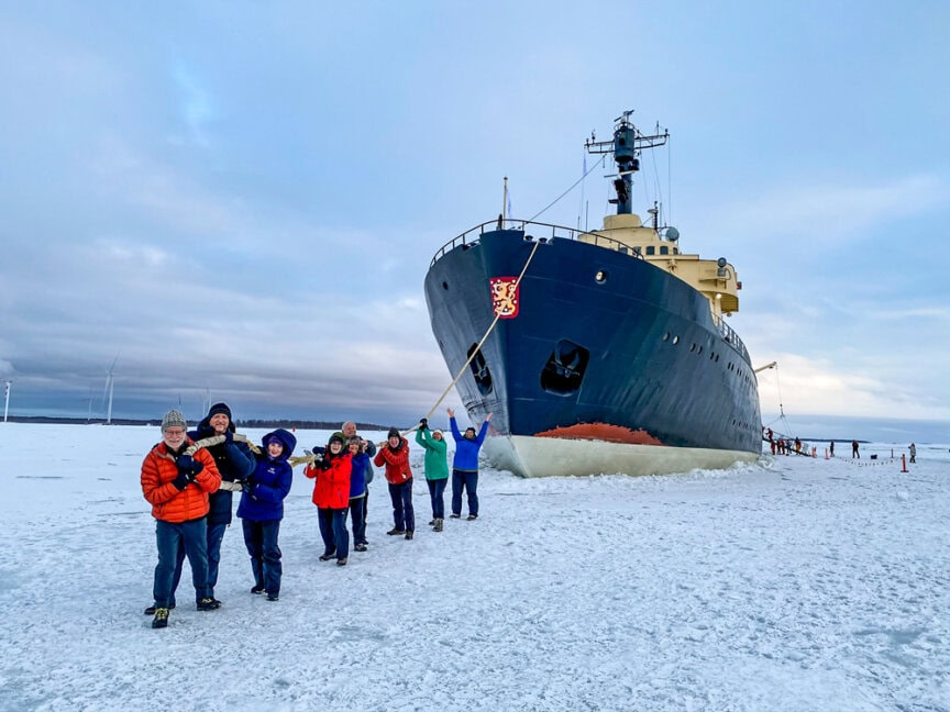 A group of people pose on an ice-covered landscape in front of a large ship, partially embedded in the ice. Some are holding cameras and dressed in colorful winter jackets, capturing their adventures. This scene embodies the excitement of Northern Finland travel.