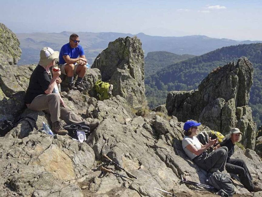 Four people rest on a rocky mountain peak, backpacks and hiking poles by their sides, taking in the panoramic view of distant hills under a clear sky. Their journey is part of the Great Carpathian Traverse, echoing the call of their Carpathian roots.