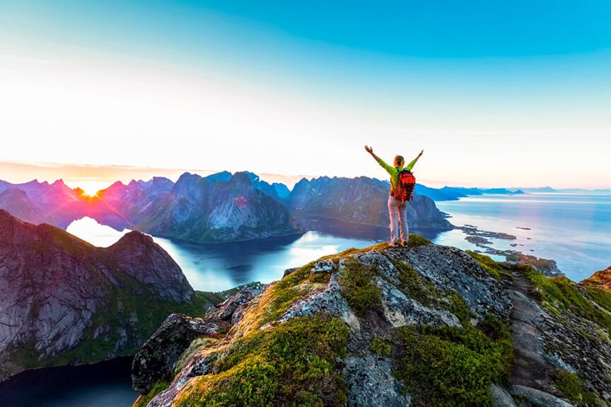 A traveler with a backpack raises their arms while standing on a cliff overlooking a sunset-lit fjord and mountains in Europe.