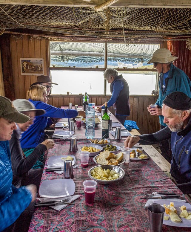 People are seated around a dining table with plates, cups, and assorted food. Two individuals are standing, one serving himself food. The room's wooden walls and overhead nets give it a rustic charm, reminiscent of adventures in Ushuaia at the End of the World.