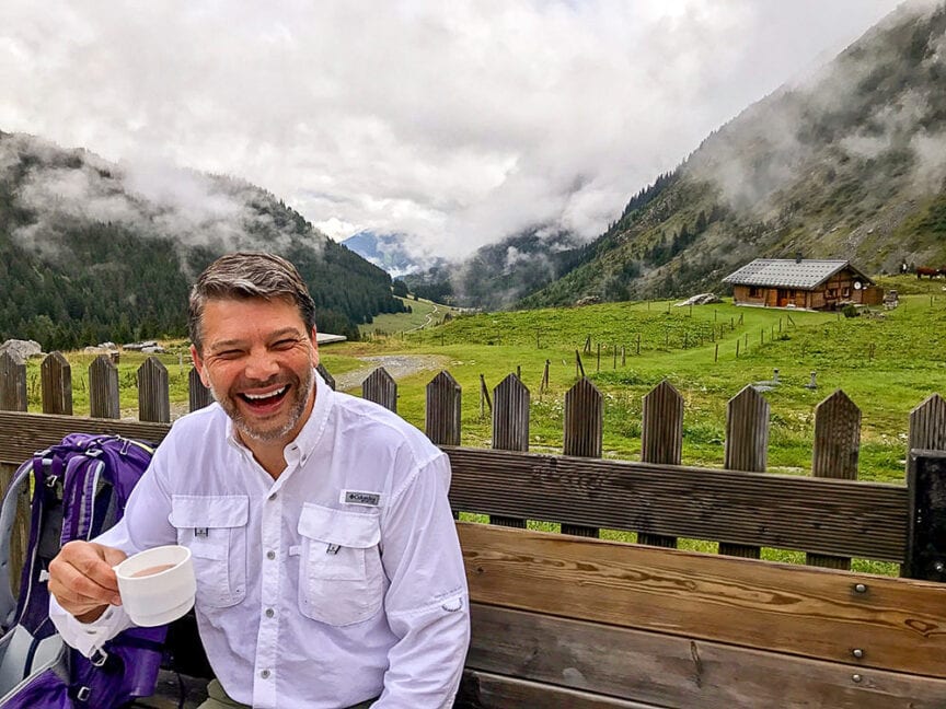 A man with a beard and mustache sits on a bench, smiling and holding a cup of coffee. In the background is a scenic valley with mountains, a cabin, and clouds amidst green fields, reminiscent of the landscapes along the Tour du Mont Blanc hiking trail.
