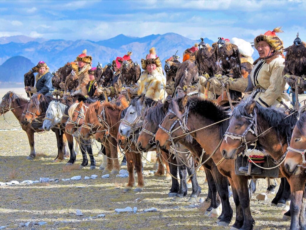 A group of hunters in traditional Mongolian attire sit on horseback, each holding a trained golden eagle, with mountains in the background.