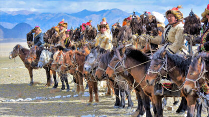 A group of hunters in traditional Mongolian attire sit on horseback, each holding a trained golden eagle, with mountains in the background.