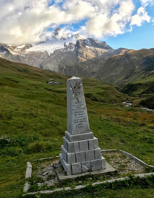 A stone monument with inscriptions stands in a grassy landscape, set against the backdrop of snow-capped peaks partially veiled by clouds, reminiscent of views along the Tour du Mont Blanc hiking trail.