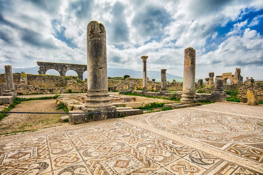 Ancient Roman ruins with standing columns and mosaic floor patterns evoke a timeless urban antiquity under a partly cloudy sky.