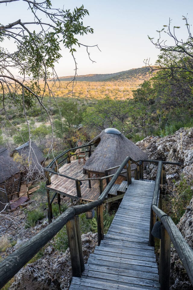A wooden staircase leads down to a rustic hut with a thatched roof in Namibia's hilly, dry landscape, where sparse vegetation hints at the region's diverse wildlife.