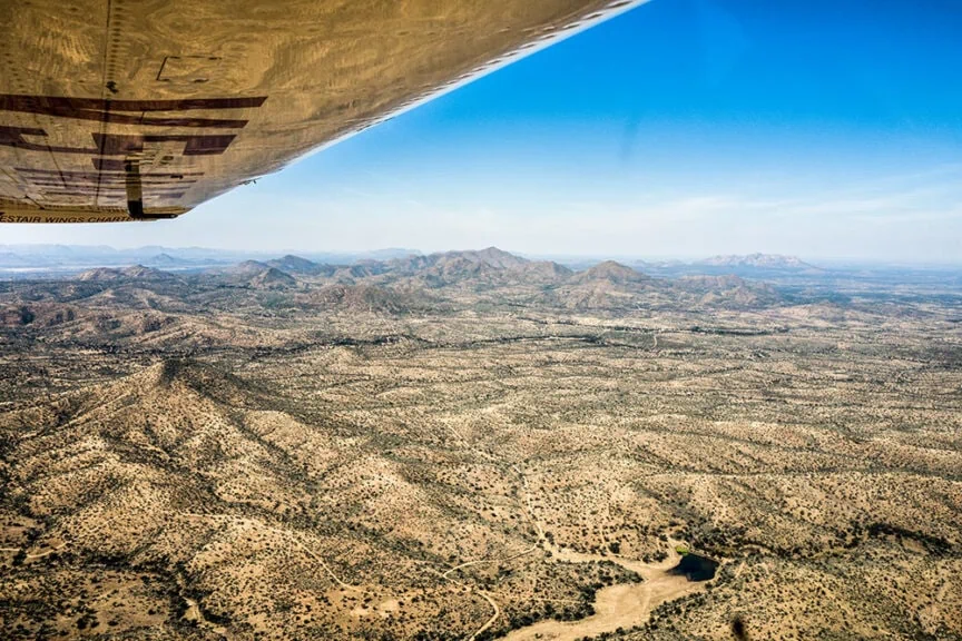 View from a small aircraft showing an expansive Namibian desert landscape with distant mountain ranges under a clear blue sky.