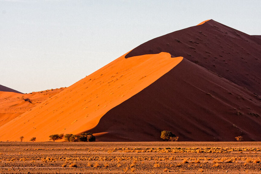 A sunlit desert sand dune with a steep slope casts shadows on one side, with a flat plain and sparse vegetation at its base, characteristic of the stunning landscapes in Namibia.