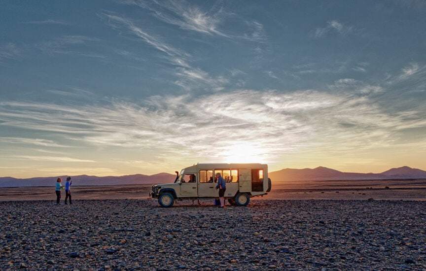 A group of people stand beside a safari vehicle in the rocky desert landscape of Namibia at sunset, with mountains in the distance and a cloudy sky overhead, capturing the essence of wild landscapes.