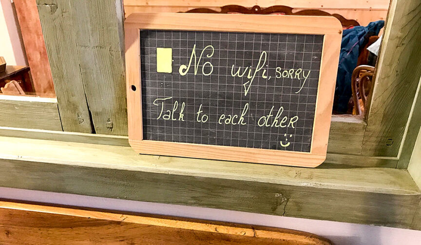 Small chalkboard sign in a wooden frame reads, "No wifi, sorry. Talk to each other," with a smiley face at the end. Perfect for those returning from a Tour du Mont Blanc hike, it's displayed on a window ledge in a rustic setting.