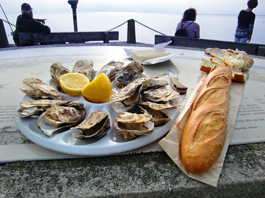 A plate of opened oysters with lemon wedges, a long baguette, and a partial view of a body of water with people sitting and looking out in the background—truly food travel at its finest.