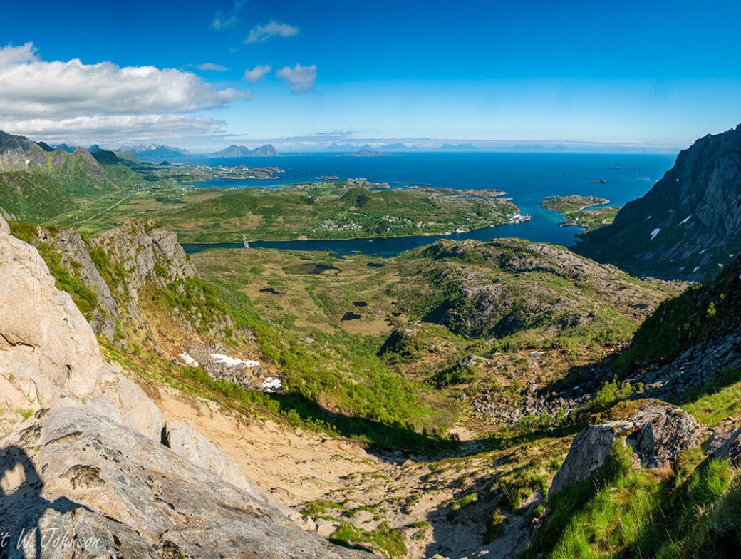 Panoramic view of a Norwegian landscape featuring a valley, rocky cliffs, scattered greenery, a coastal town, and blue waters under a clear sky with some clouds. Ideal for adventures near the Arctic Circle.