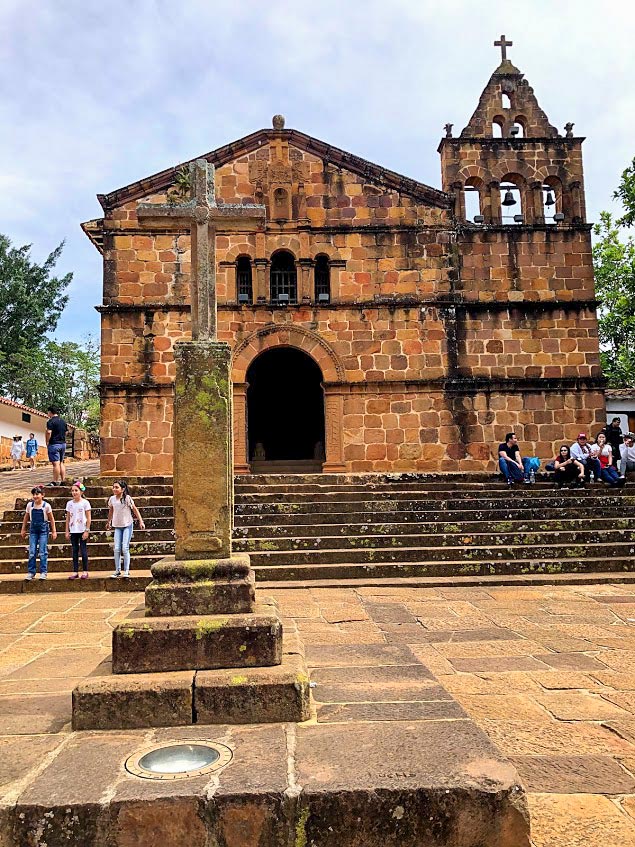 The stone church with a cross in front features a weathered exterior and steps leading to its entrance. A small group of people is seated on the steps, with others standing nearby, creating the perfect snapshot of amazing Colombia experiences. Trees are visible in the background, adding to the charm.
