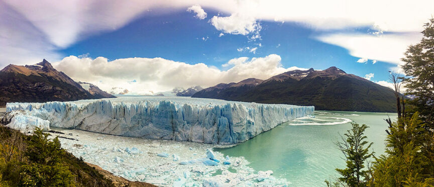 A panoramic view of a glacier in a mountainous landscape within Patagonia's renowned national parks, with a clear sky above and chunks of ice floating in the greenish water in the foreground—perfect for those who love hikes.