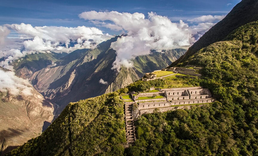 Aerial view of Machu Picchu set against a backdrop of lush green mountains and clouds. The stone ruins of the ancient Incan city in Peru are spread across the mountaintop, showcasing a breathtaking urban antiquity.