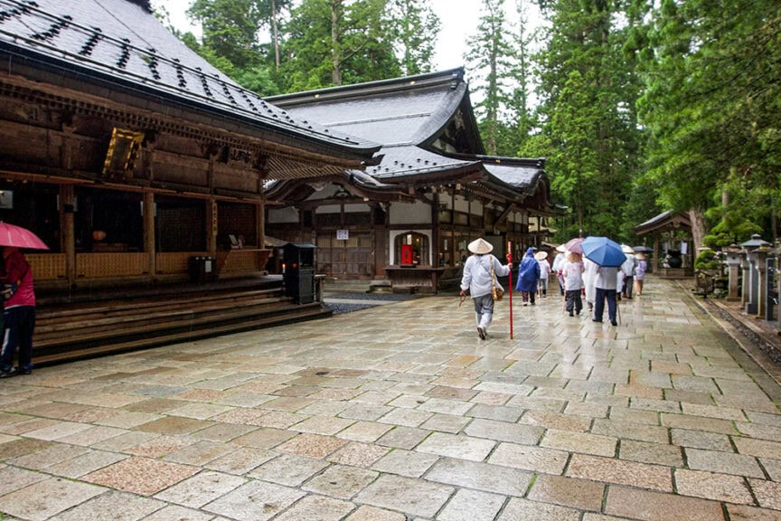 People exploring the lush, rainy setting of Koyasan walk with umbrellas and traditional hats near a wooden temple, experiencing the essence of Ancient Japanese Culture.