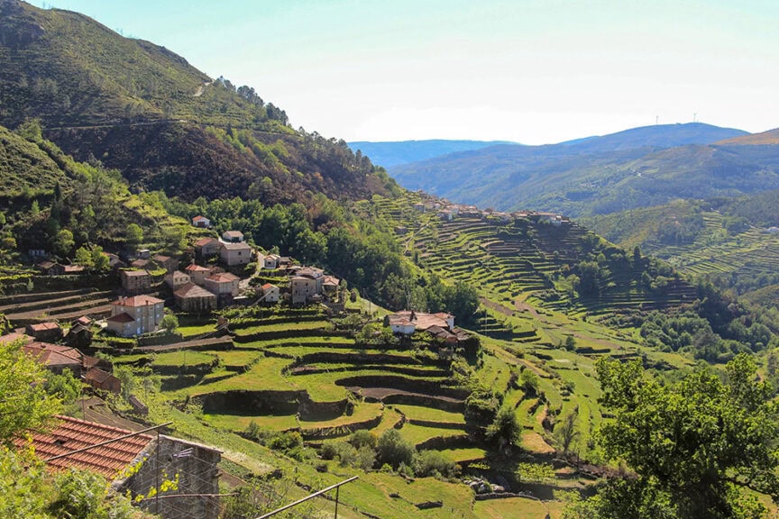 Hillside village with terraced green fields, scattered houses, and a distant mountain range under a clear sky, embodying the simple pleasures of rural Portugal.