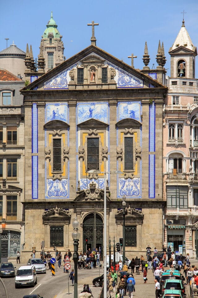 A historic blue and yellow tiled church facade with a cross on top, nestled among older buildings in rural Portugal, and overlooking a busy street where the simple pleasures of daily life unfold amidst people and cars.