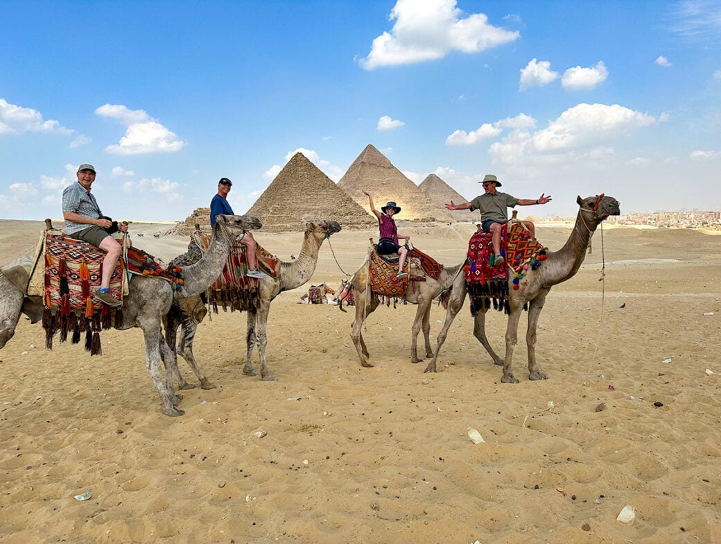 Four people embark on Desert Adventures, riding camels in front of the pyramids under a blue sky with scattered clouds, capturing the essence of Egypt's timeless landscape.