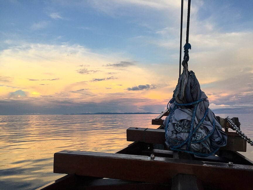 A boat's bow with rigging is shown facing a calm, reflective body of water during a tranquil sunset. The sky displays a gradient of blue, pink, and orange hues with distant clouds on the horizon, evoking the serene beauty often sought in wild frontier adventures.