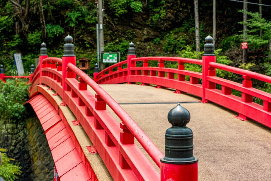 A curved red bridge with black finials, reminiscent of ancient Japanese culture, crosses a wooded area. Trees and green signboards are visible in the background, evoking the serene beauty of Koyasan.